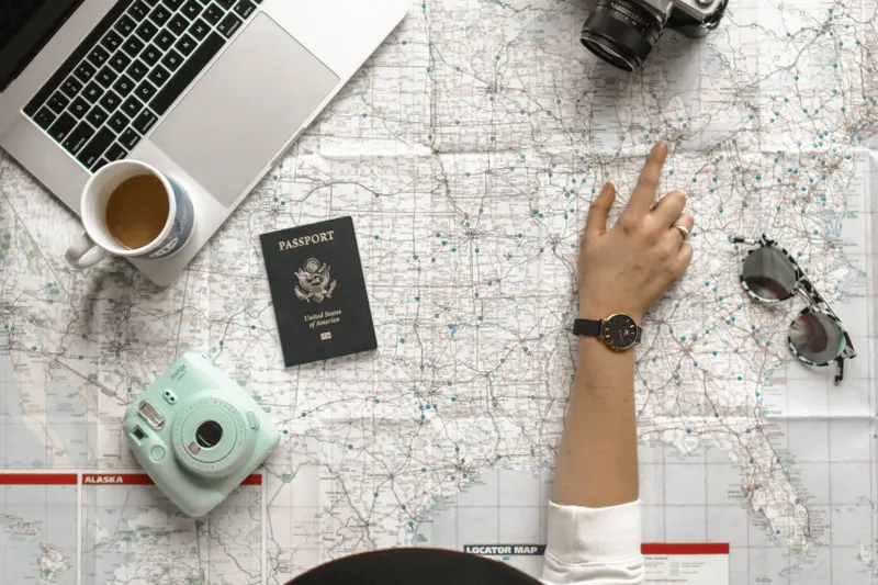 A hand pointing to a map beside a laptop and travel-planning essentials